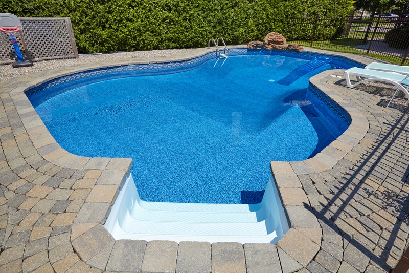 A vinyl swimming pool with crystal blue water is nestled in a backyard with a stone patio.