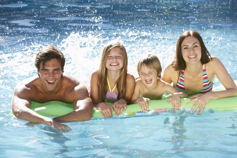 A family shares a float and smiles at the camera as they splash in their swimming pool.