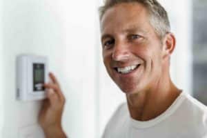 A smiling older gentleman wearing a white shirt adjusts his HVAC system using the thermostat in his home.