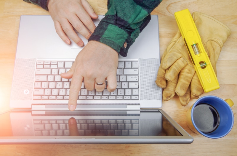 A contractor manages online promotion for his contractor business at a laptop beside his work gloves, bubble level, and coffee.