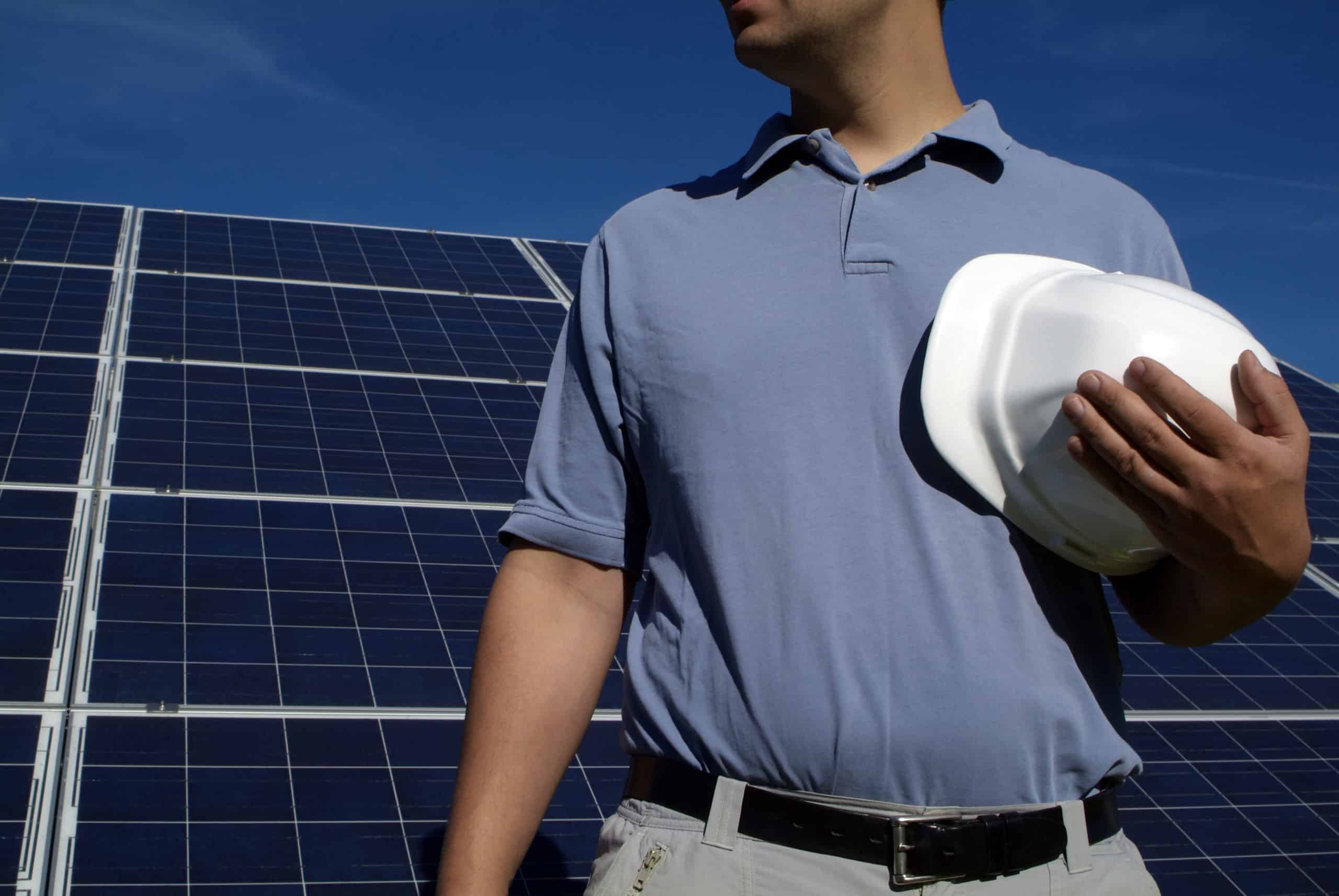 male contractor holding a white hard hat stands in front of large solar panels