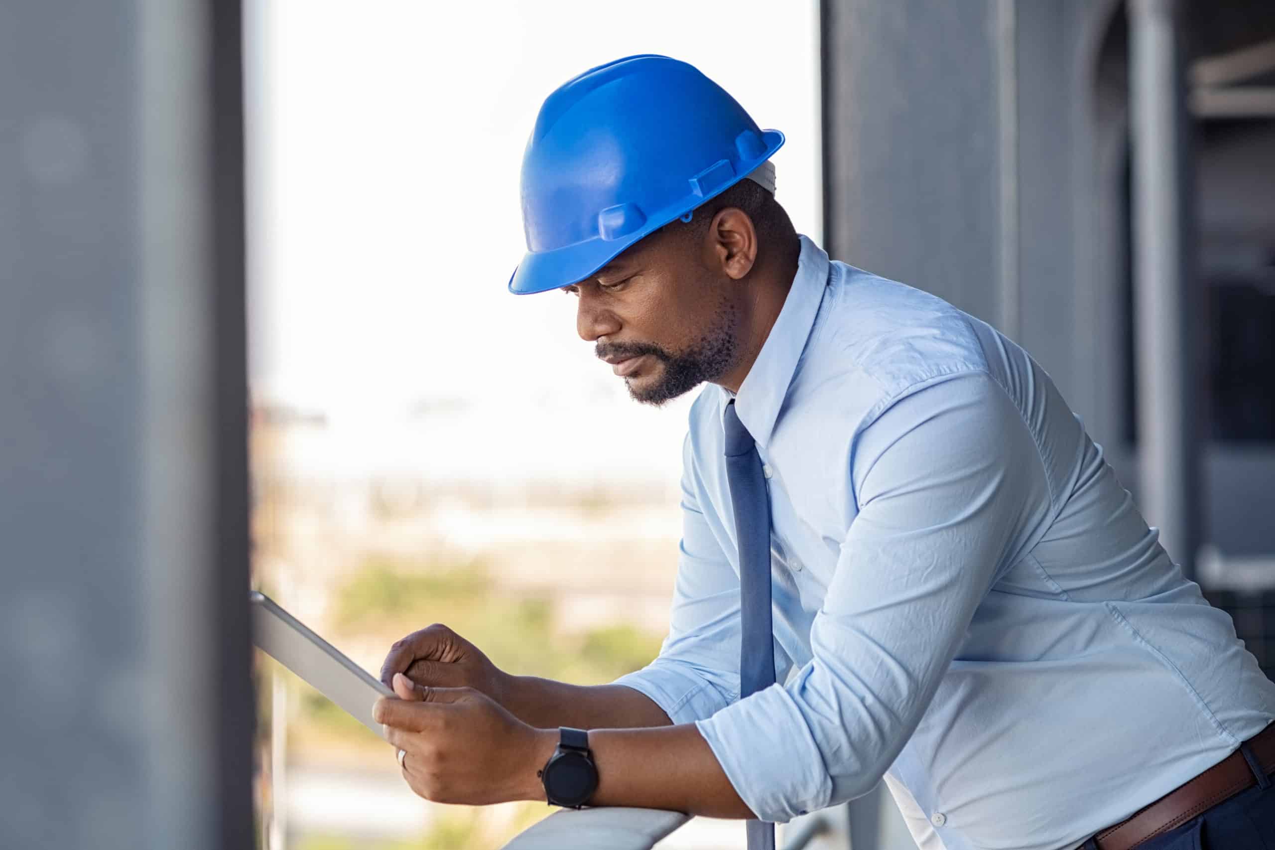 A general contractor wearing a blue hardhat checks his professional email communication on his tablet.