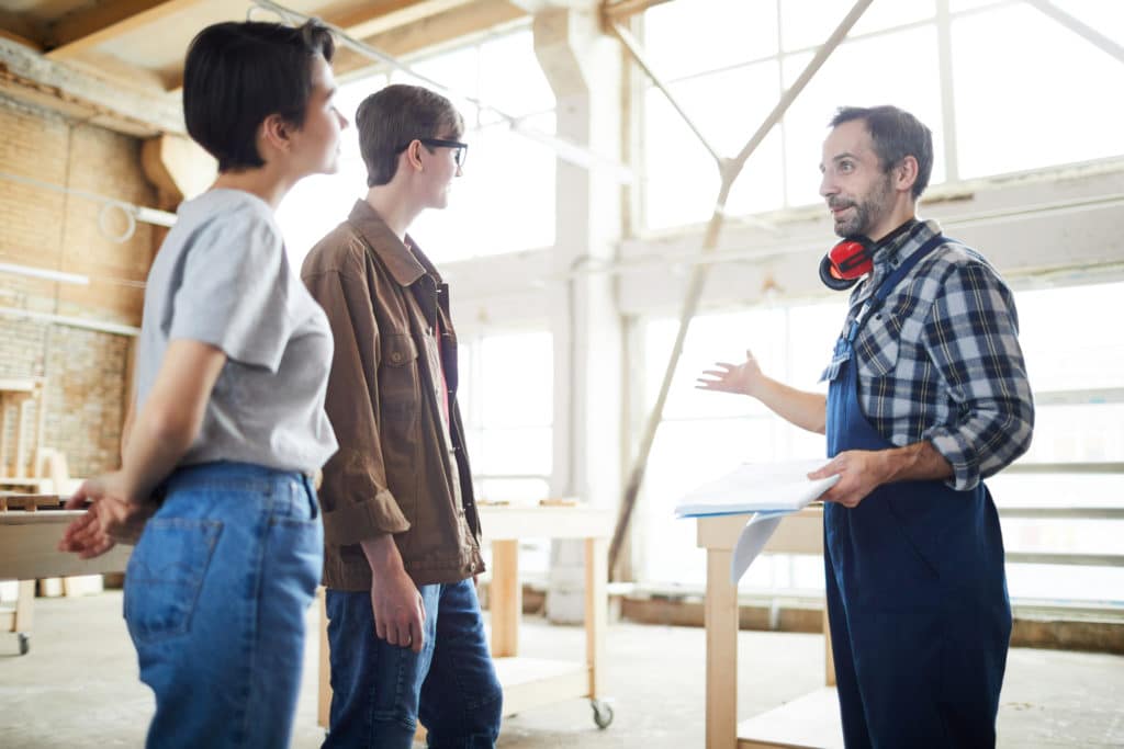 A general contractor business owner explains a building project to two clients on the building site. They are in the background while there is scaffolding visible in the background.