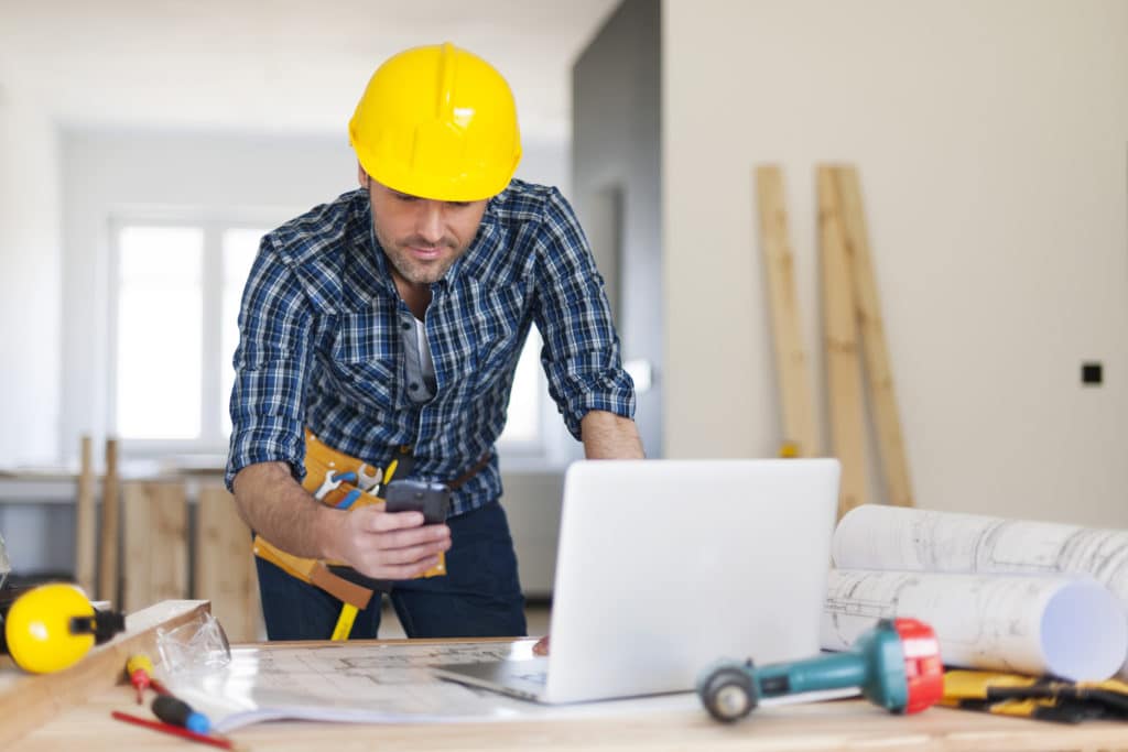 A general contractor business owner checks his computer screen while on a break during a project. His table is covered with tools and plans.