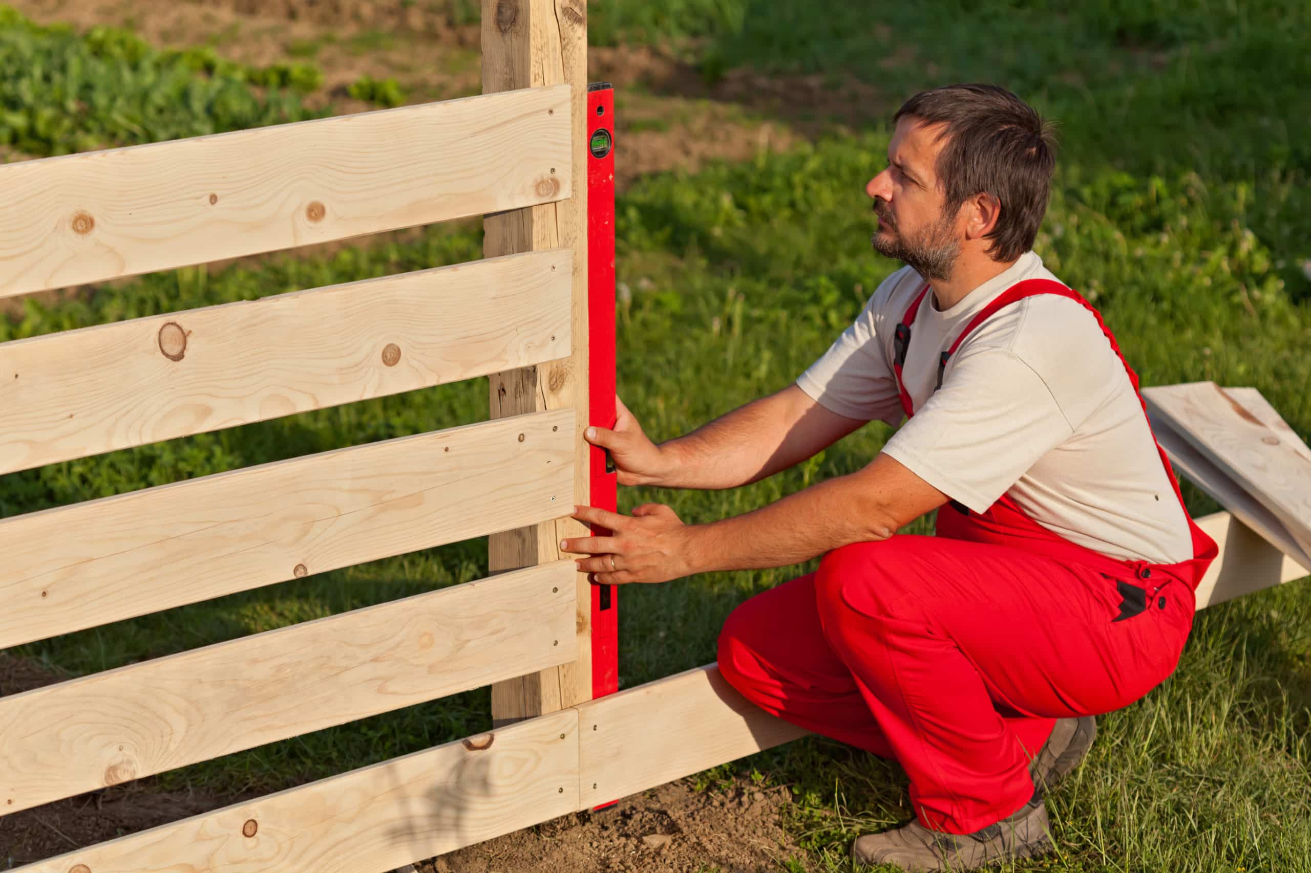 A fencing and decking business contractor in red overalls uses a level to make sure his fence is square.