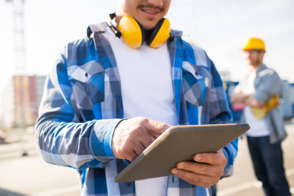 A deck and patio business owner wearing a blue plaid shirt and yellow hearing protection down around his neck holds a tablet to monitor his website.