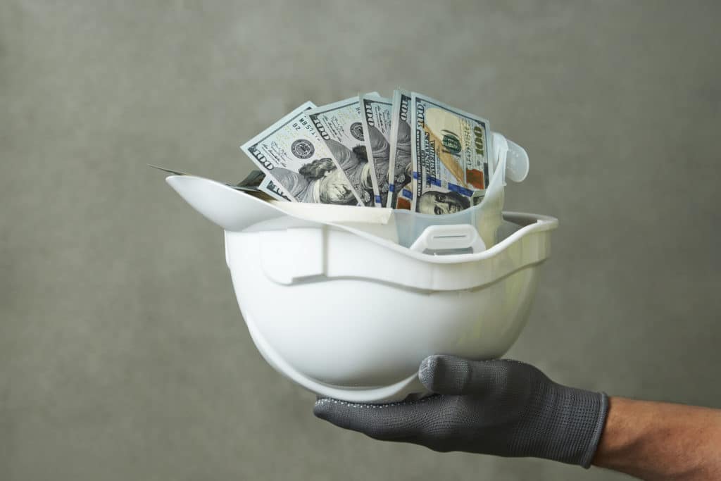 A white hard hat filled with cash sits in the gloved hand of a construction worker. 