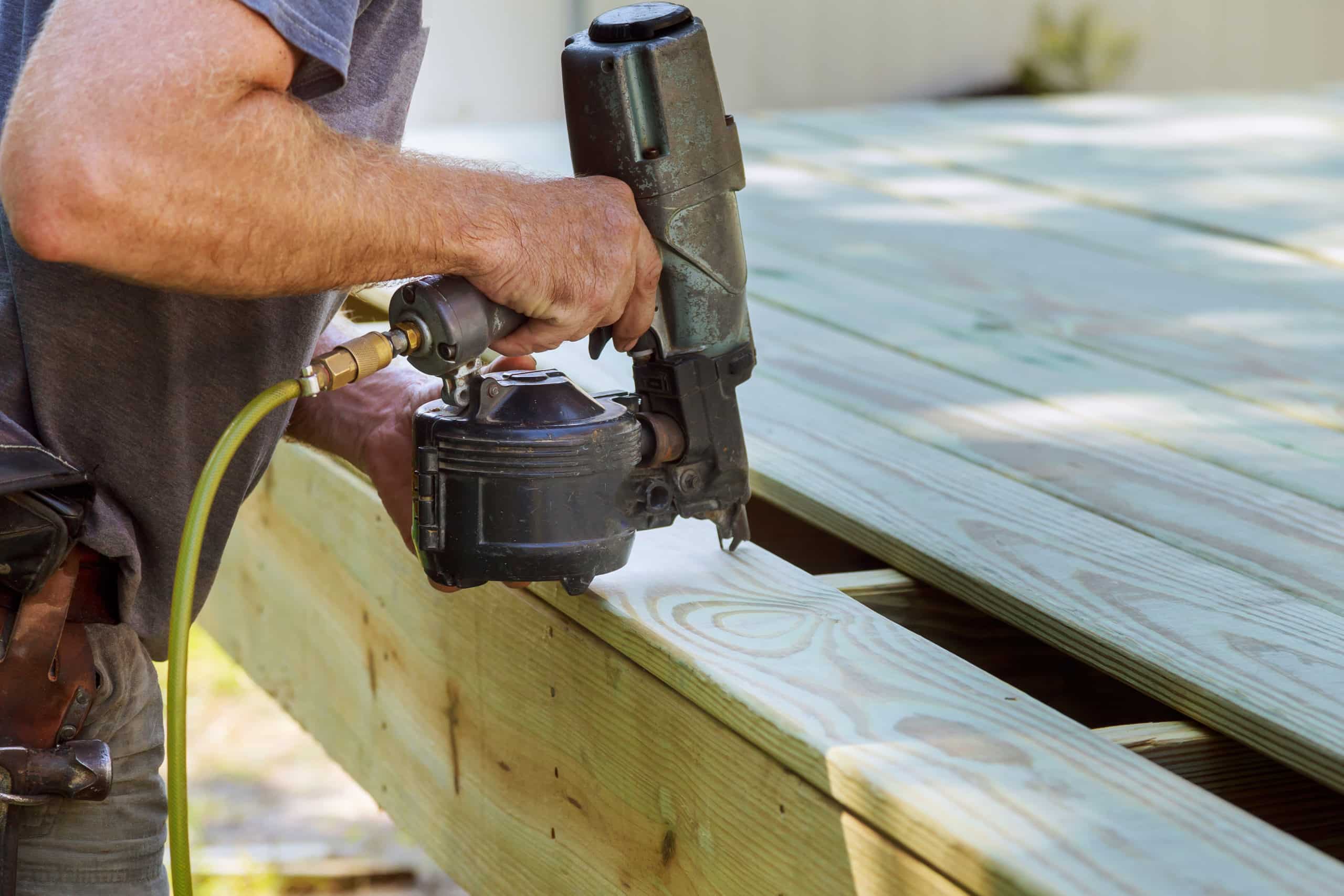 A fencing and decking business contractor operates a pneumatic nail gun to finish building a new deck.