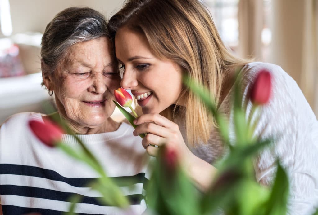 An elderly woman smells red tulips with her grown granddaughter. They may consider contractor financing for an Accessory Dwelling Unit.