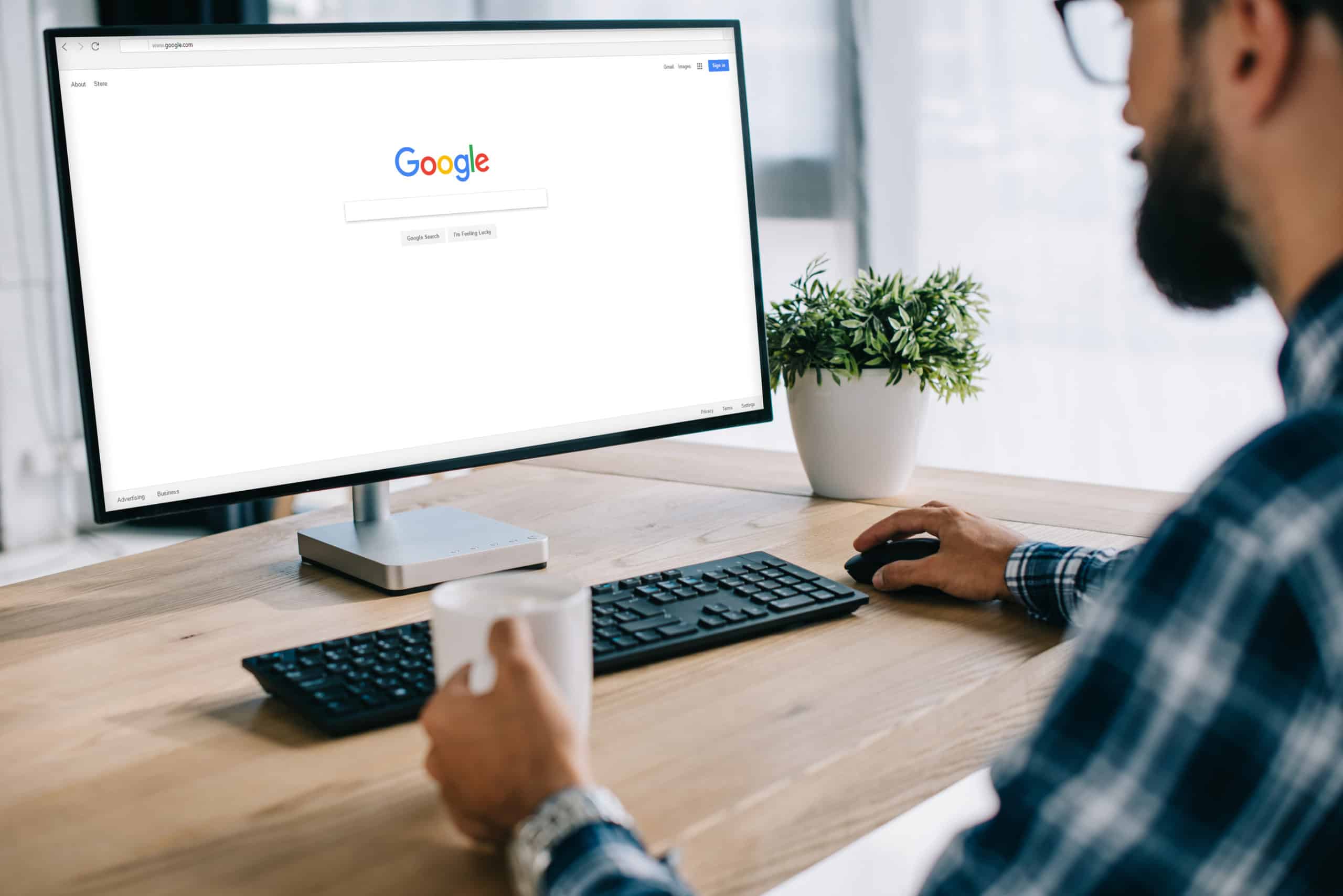 A bearded man in blue flannel holds a coffee cup in his left hand and clicks on the Google search page with his left. He is going to research Google rankings for fencing and decking businesses.