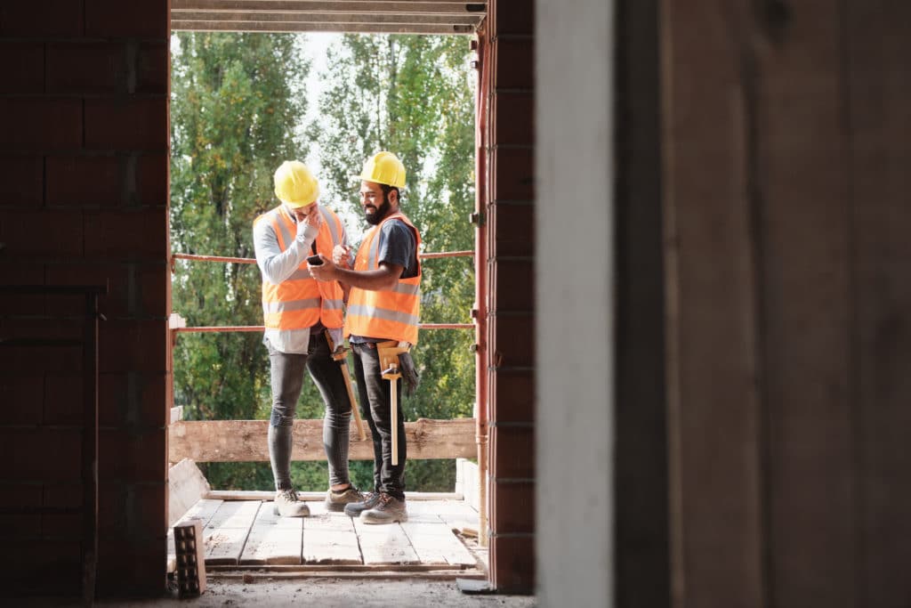 Two contractors laugh at a cell phone. They are framed by a door and are standing on a deck.