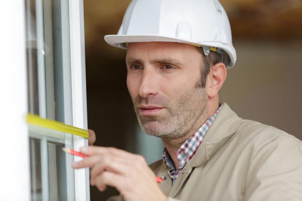 Hiring a door and window contractor with the right qualifications is important. The window installer wearing a white hard hat measures the window's edge as he installs it. 