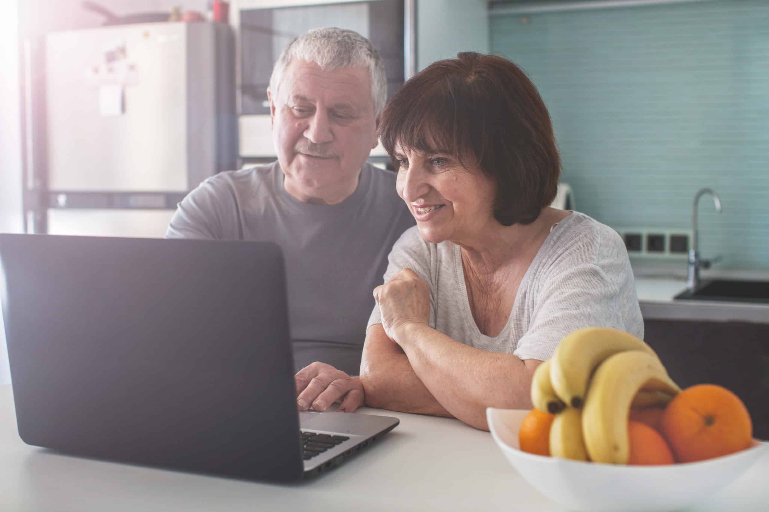 An older couple sits at their kitchen counter looking at a laptop computer to check reviews and references for a fencing and decking contractor.