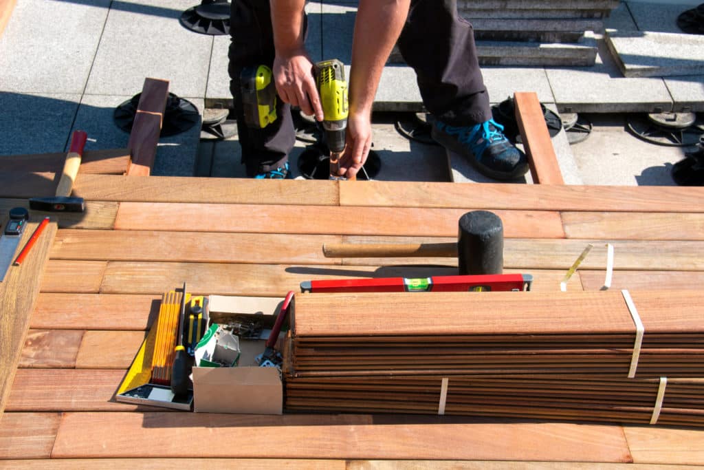 A decking contractor builds a perfect deck which is why he has perfect online reviews. His materials and tools are stacked in front of him as he drills in another board.