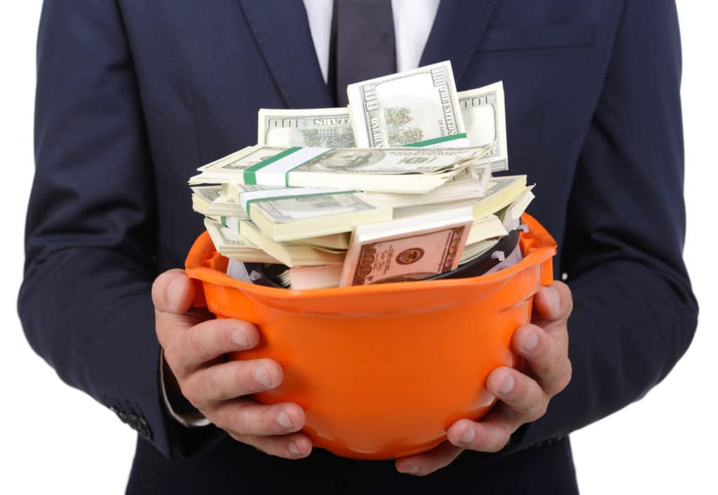 A man wearing a business suit holds out an orange hard hat full of cash.
