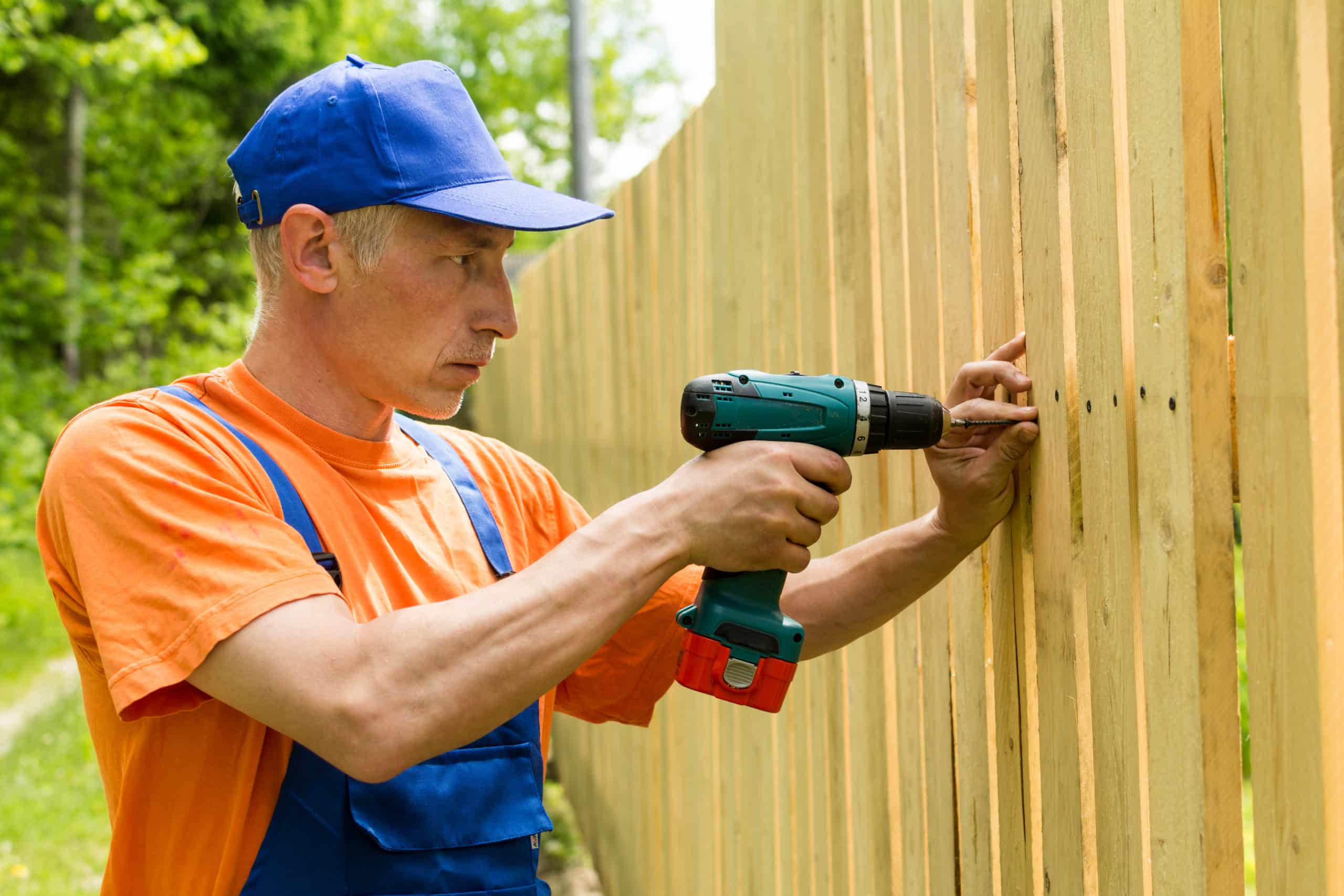 A fencing and decking contractor can keep costs down by paying close attention to his fence build. The contractor is wearing an orange shirt with a blue hat and coveralls. He's drilling into a wooden fence with a cordless drill.