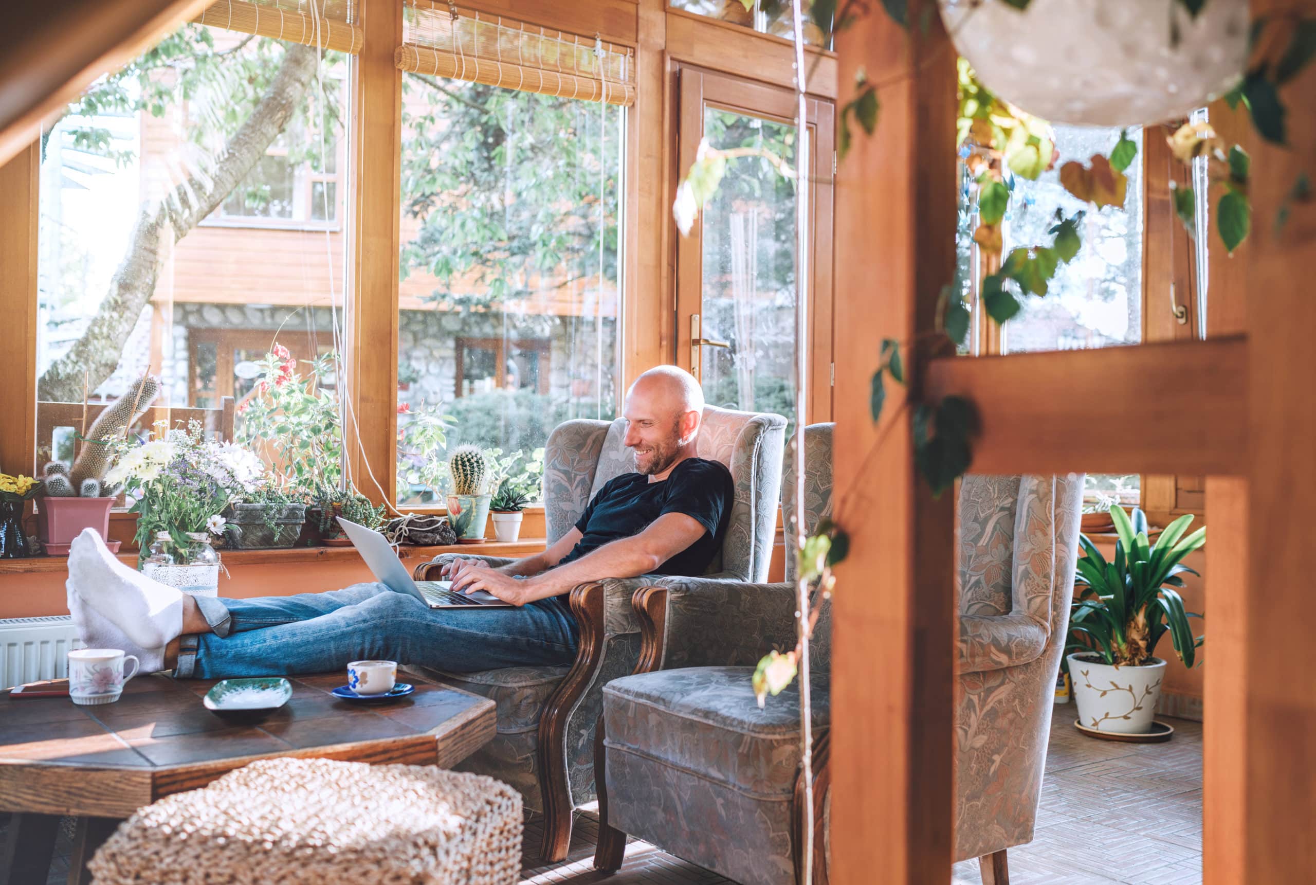 A man wearing a black shirt lounges in his beautiful sunroom with natural wood beams while building a website on his laptop for his job as a sunroom builder.