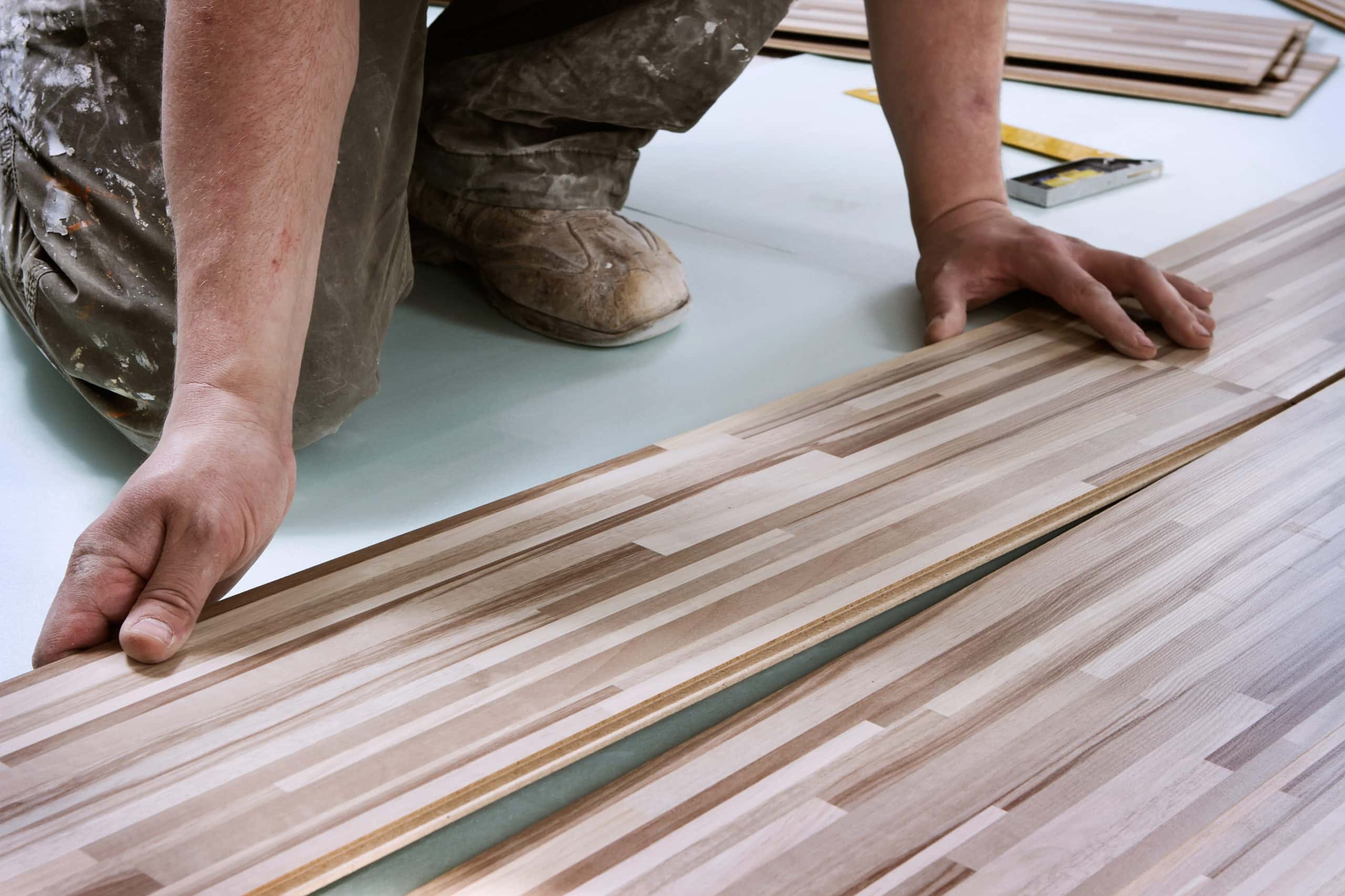 A home improvement contractor installs wooden floor planks while on his knees. We can see his hands and work boots as he kneels.