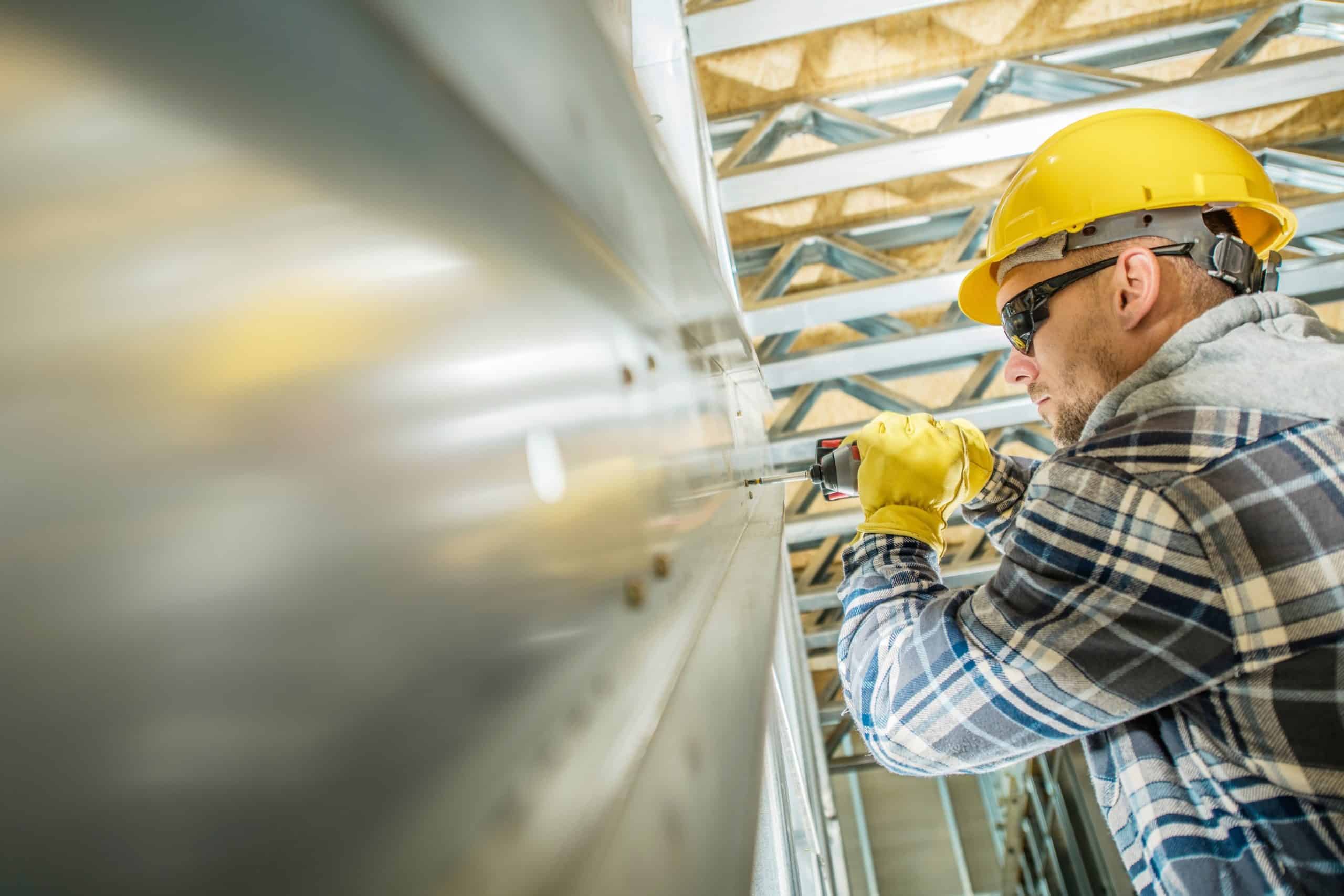 A steel building contractor drills into a wall as he dominates the competition by building the best. He is wearing flannel and a yellow hard hat.