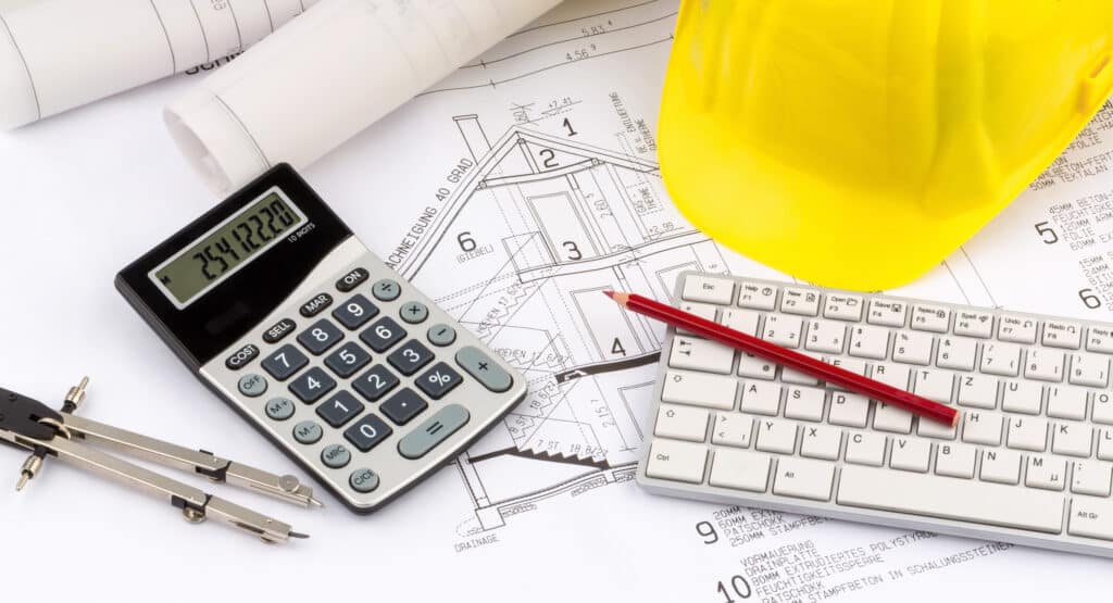 A calculator, keyboard, and yellow hard hat sit atop some blueprints where a home improvement contractor has been calculating costs in order to dominate the competition within your market. 