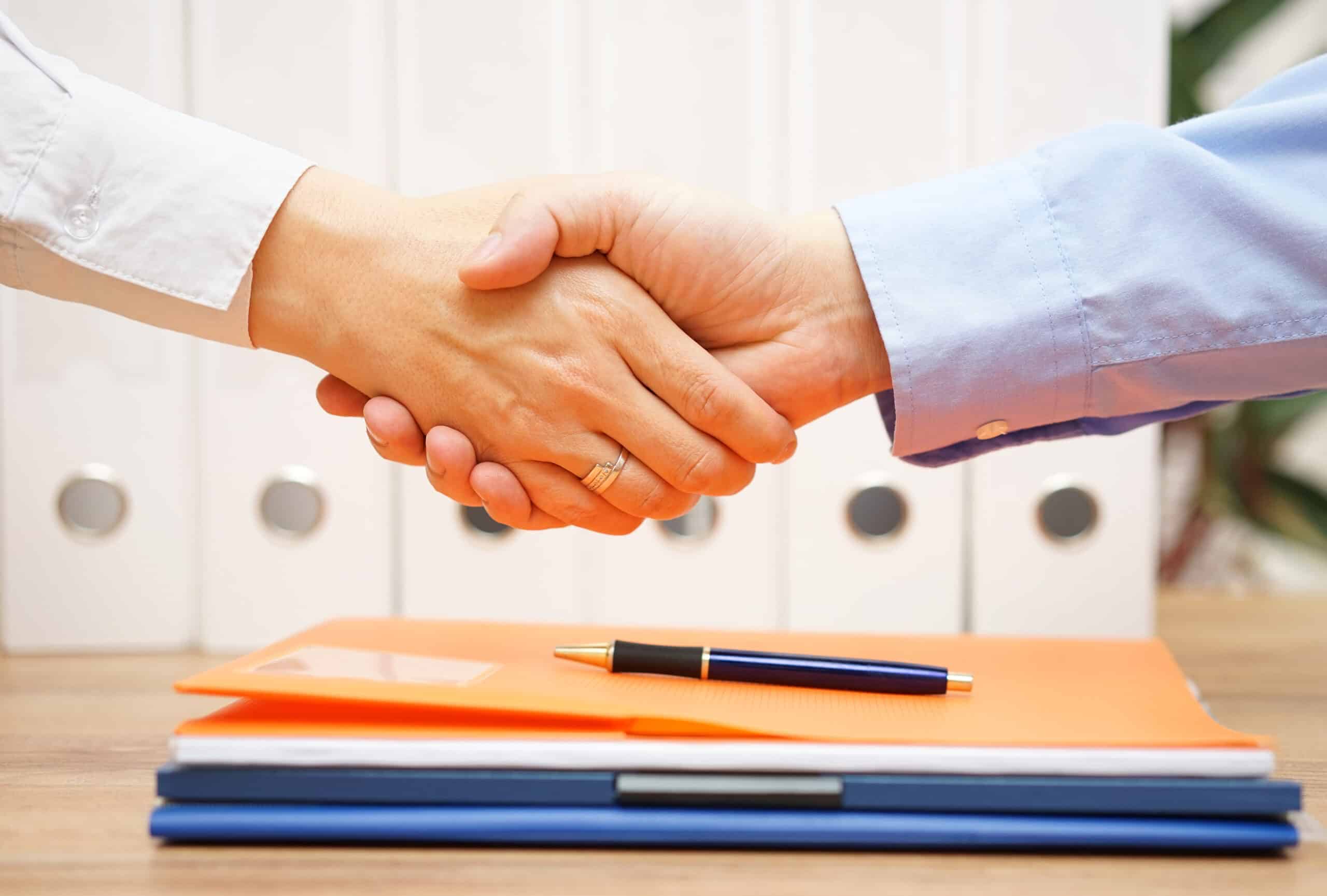 Two professionals shake hands over a stack of documents you should always provide your sunroom clients that's sitting on a table.