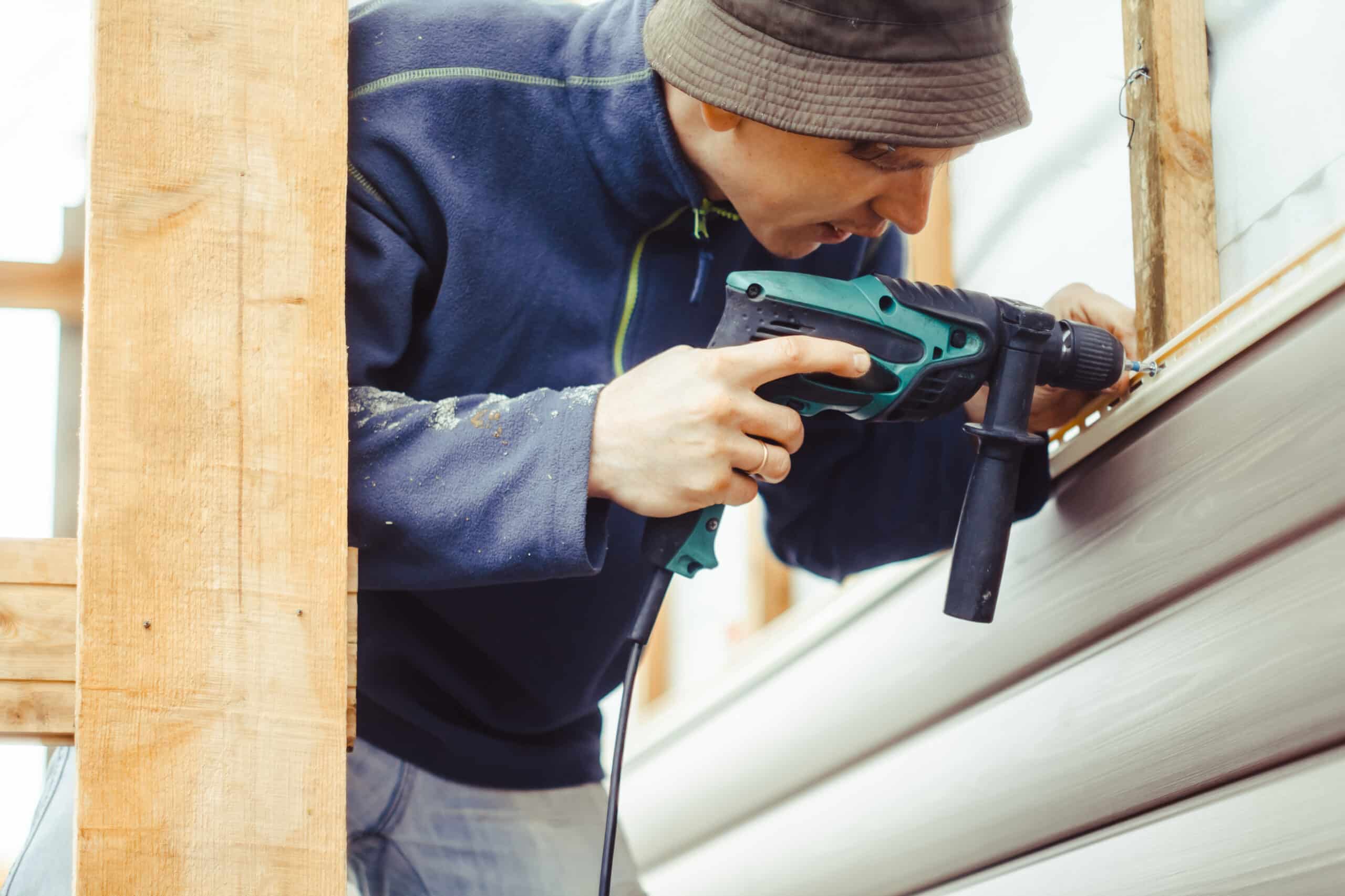 A siding contractor dressed for cold weather installs new beige siding with a nail gun. He is dedicated to hard work in order to win more customers.