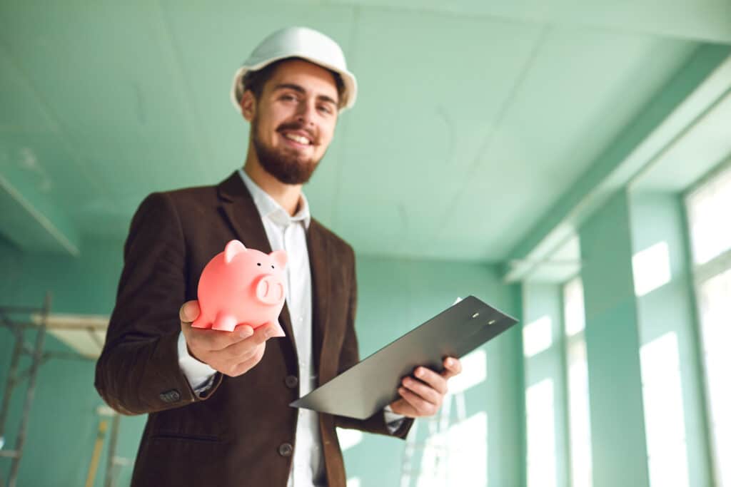 A sunroom builder wearing a sports jacket and a hard hat holds a piggy bank in one hand and a clipboard in his other. He stands in a green sunroom.
