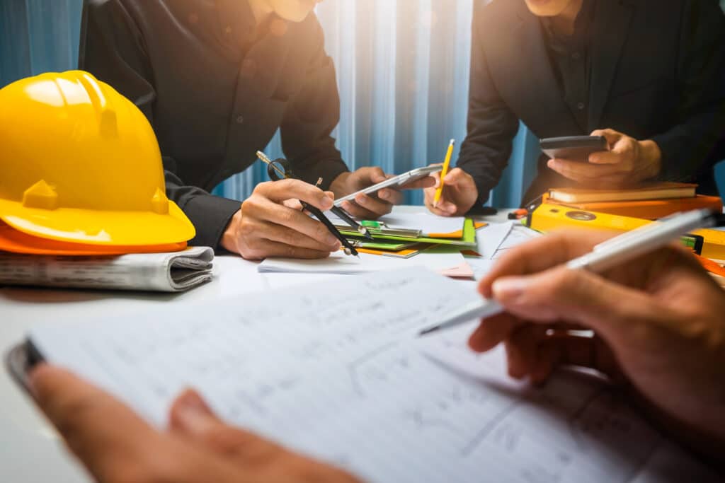 A team of sunroom builders guide each other to better business practices as they discuss business over building plans. Each person is holding a pen, and a hard hat sits atop the table.