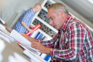A home renovation contractor reviews the documentation you should always provide your home renovation clients. He's wearing a red plaid shirt and leaning over a table covered with paperwork while his associate organizes a shelf in the background.