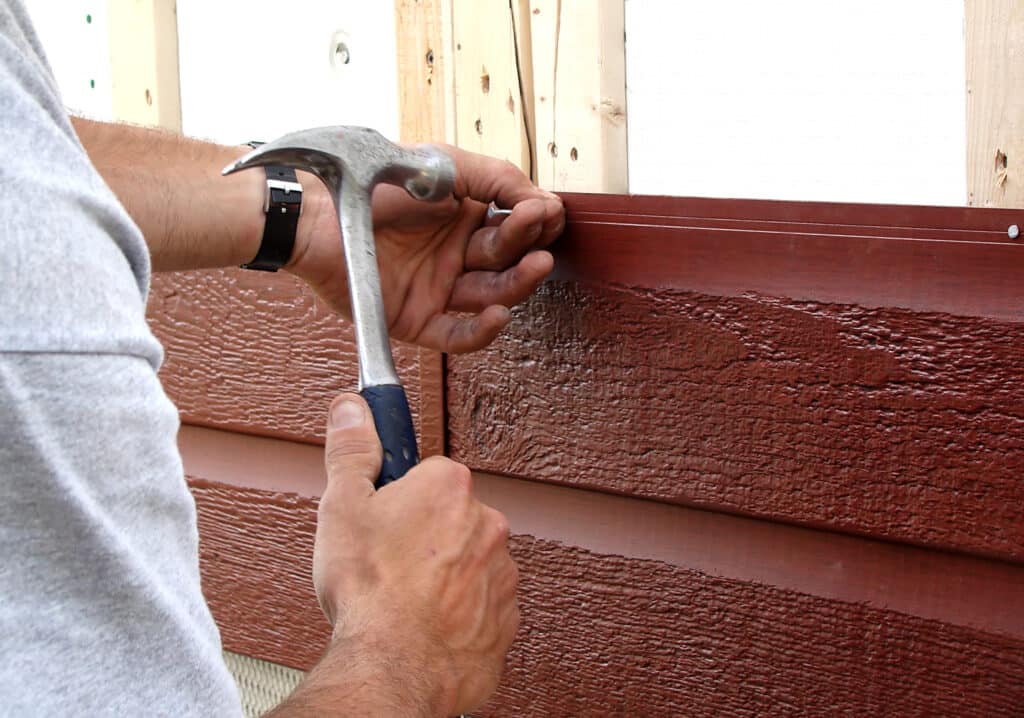 A gutter and siding contractor installs new, dark red siding with a hammer and nail. 