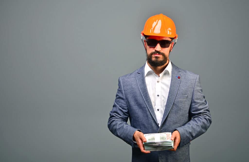A contractor is wearing an orange hard hat, dark sunglasses, and a gray sport jacket while holding a stack of cash.