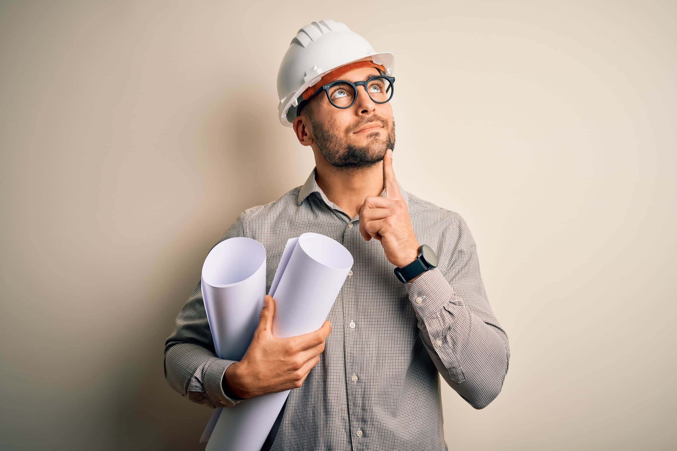 A contractor holding building plans and wearing a white hard had looks as if he's thinking and pondering the benefits of financing through HFS Financial.