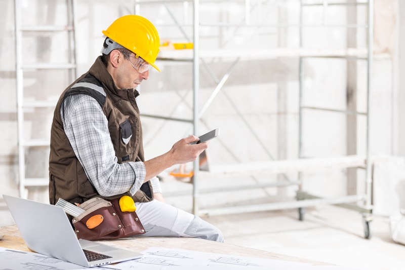 A general contractor wearing a yellow hardhat looks on his phone for ways to become the top financing contractor.